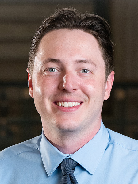 A white man with short dark hair wearing a baby blue button-down and tie