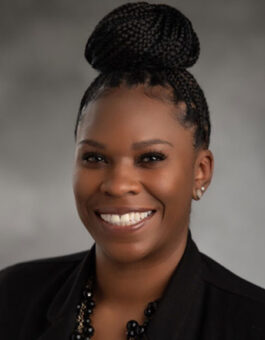 A black woman with box braids in a bun wearing a chunky black necklace, earrings and a black blazer.