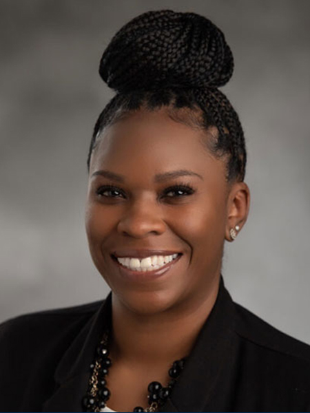 A black woman with box braids in a bun wearing a chunky black necklace, earrings and a black blazer.