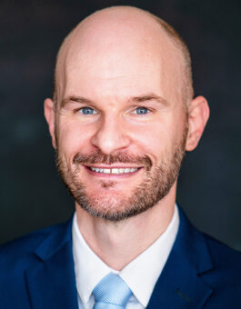 A white man with a shaved head and beard wearing a blue suit and tie.