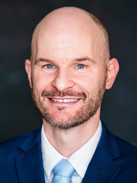 A white man with a shaved head and beard wearing a blue suit and tie.